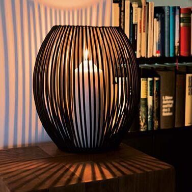 Ein Windlicht namens »Louisiana« aus Metall steht auf einem Holztisch vor einem Bücherregal. Im Inneren des Windlichts brennt eine Kerze, die warme Lichtstrahlen durch die schwarzen Gitterstäbe wirft und eine gemütliche Atmosphäre schafft.