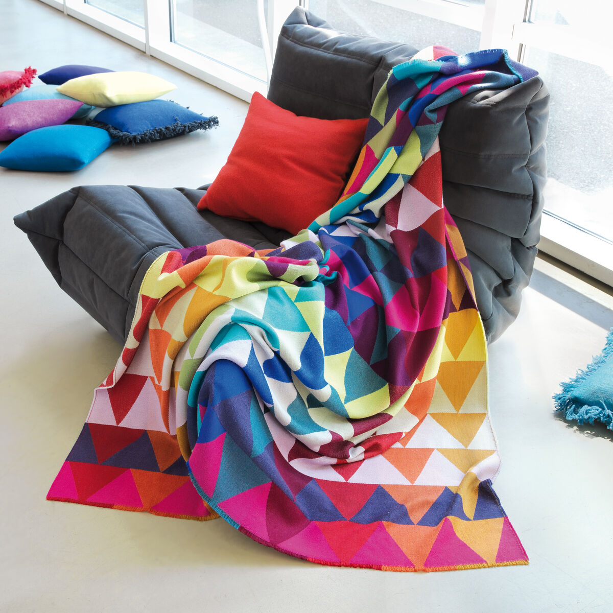Eine farbenfrohe Decke mit einem Dreiecksmuster in verschiedenen Blautönen, Rot, Gelb, Grün, Lila, Pink und Orange, drapiert über eine graue Couch vor einem modernen Fenster. Im Hintergrund sind verschiedene Kissen in Blau, Rot und Pink zu sehen.