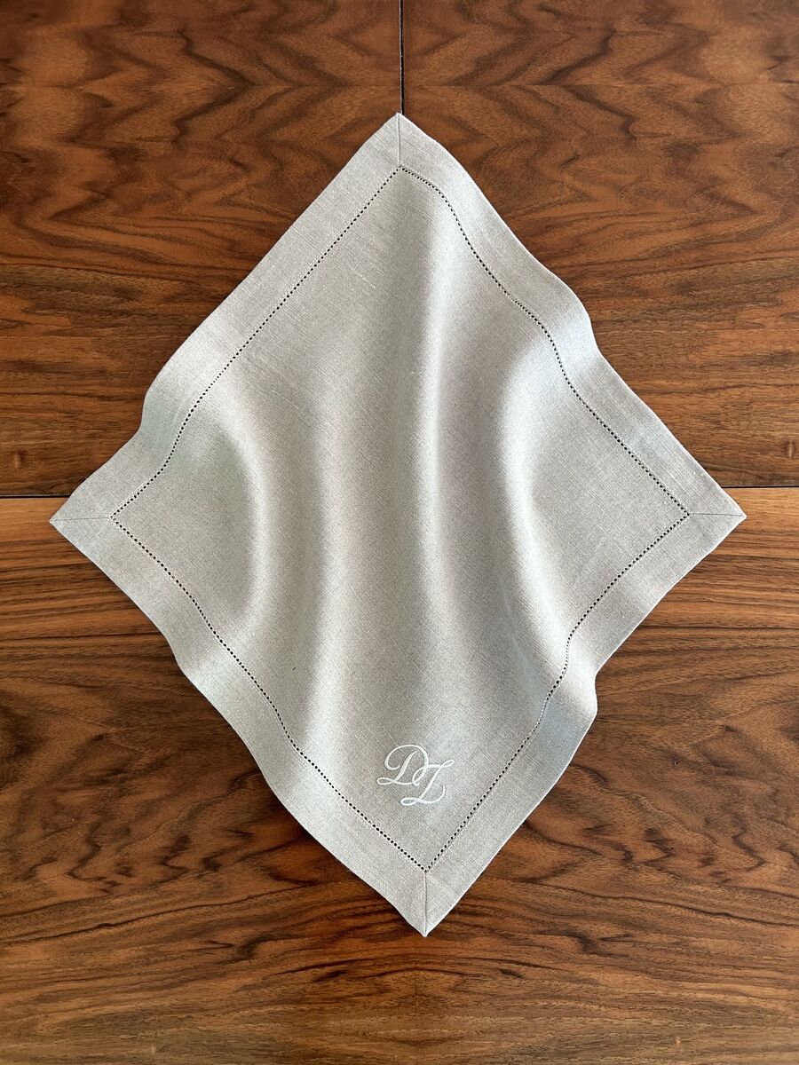 Ein quadratisches Leinen-Serviette in Naturfarbe mit Monogramm-Detail auf einer hölzernen Tischplatte.