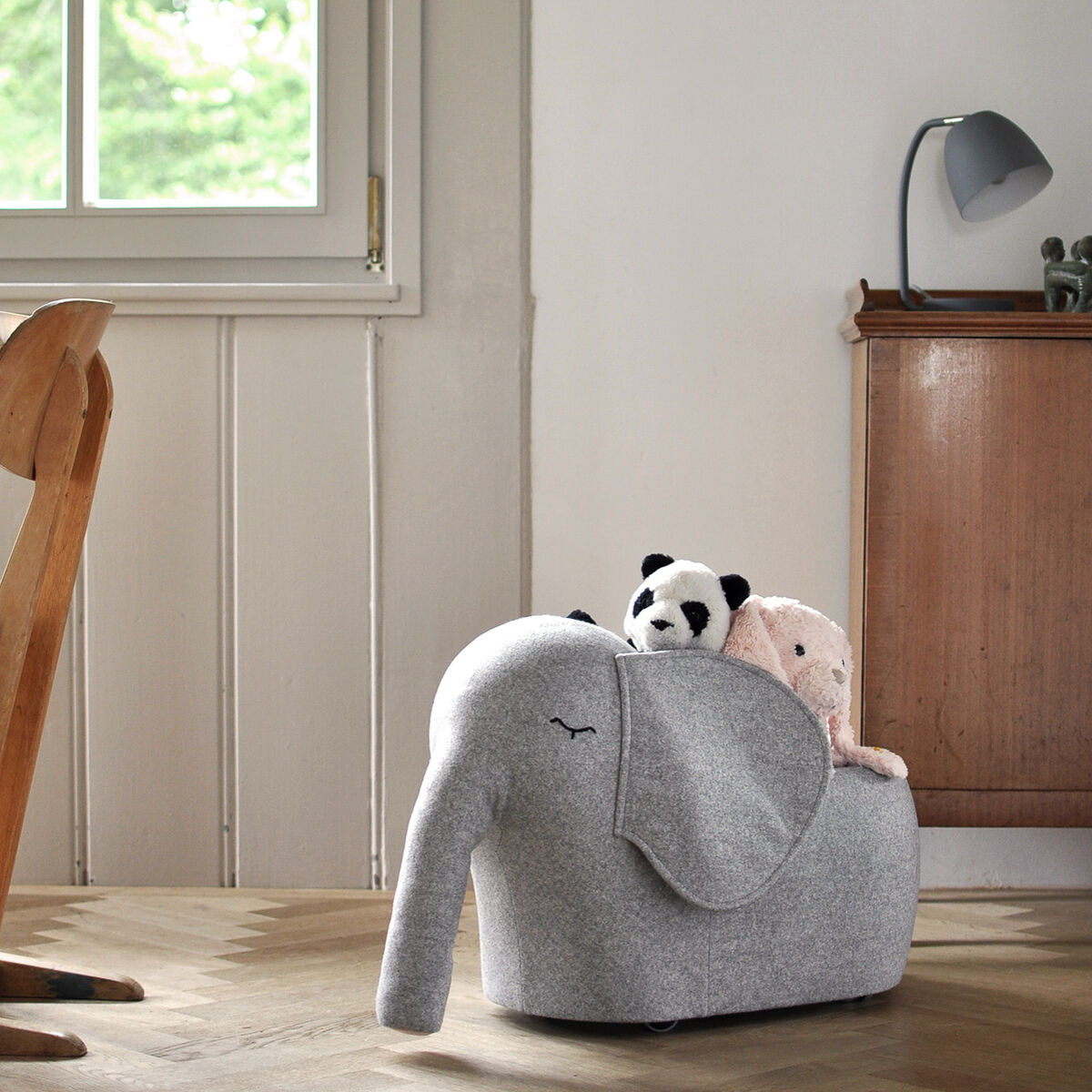 Ein grauer Rollelefant aus weichem Material steht auf einem Holzboden vor einer Kommode. Ein Panda- und ein Hasen-Kuscheltier sitzen im Rücken des Elefanten. Im Hintergrund ist ein Fenster, ein Stuhl und eine Lampe zu sehen.