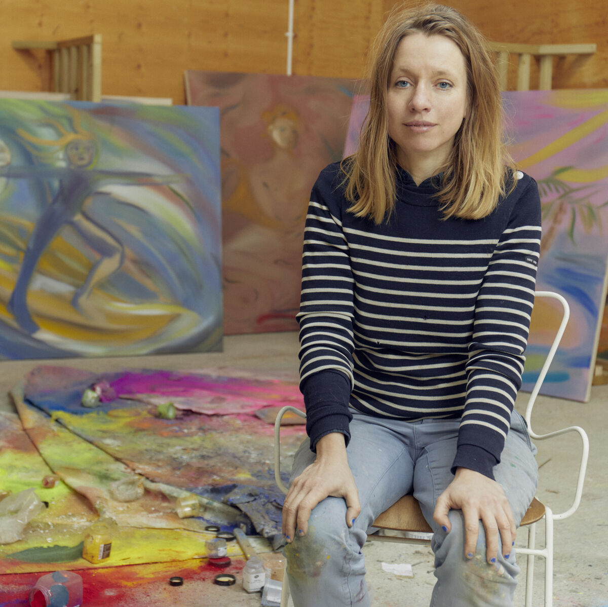 Eine Frau sitzt in einem Atelier neben mehreren farbenfrohen, abstrakten Kunstwerken auf Leinwänden. Der Boden ist mit bunten Farben und Malutensilien bedeckt.