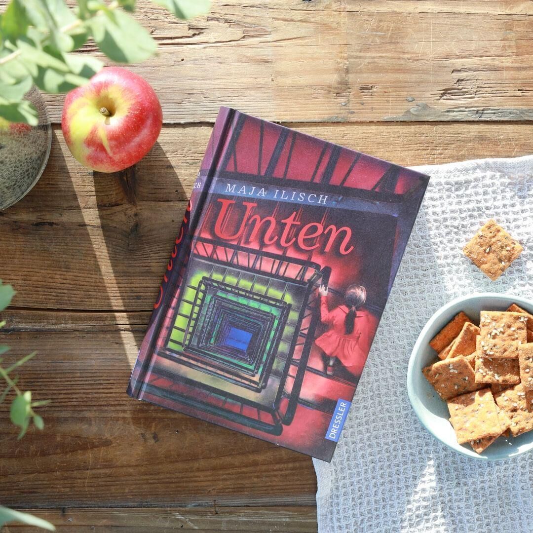 Das Bild zeigt das Buch 'Unten' von Maja Ilisch auf einem Holztisch. Neben dem Buch sind eine rote Apfel, eine kleine Pflanze und eine Schale mit Crackern, die auf einer weißen Tischdecke stehen.