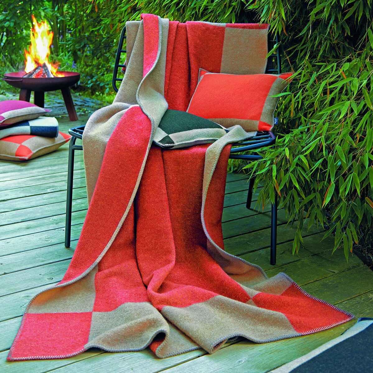 Ein gemütlicher Außenbereich mit einer schwarzen Metallstuhle, der mit einer orangefarbenen Kissenhülle und einer passenden Decke dekoriert ist. Im Hintergrund ist üppiges grünes Laub und ein brennendes Feuer zu sehen. Die Kissenhülle 'Manchester' in Orange ergänzt die herbstliche Dekoration perfekt.