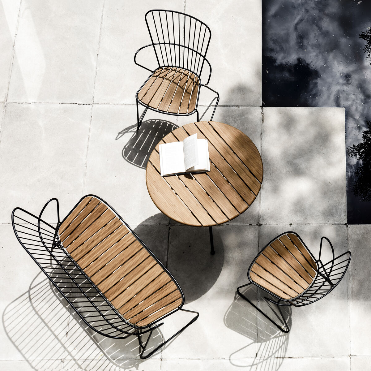 Ein Esszimmerstuhl des Modells ‚PAON‘, fotografiert in einem modernen Außenbereich. Zu sehen sind drei Stühle und eine Bank mit Holzsitz und Metallrahmen um einen runden Holztisch, platziert auf hellen Steinfliesen neben einem reflektierenden Pool.