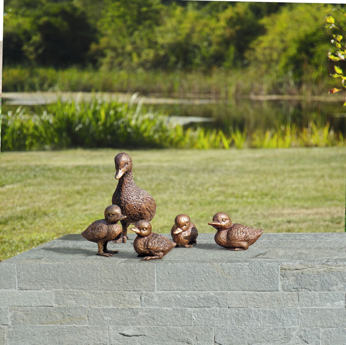 Eine Gartenskulptur der ‚Entenfamilie‘ bestehend aus einem großen und vier kleinen Enten, auf einem grauen Steinpodest im Freien, umgeben von grüner Natur und einem Teich im Hintergrund.