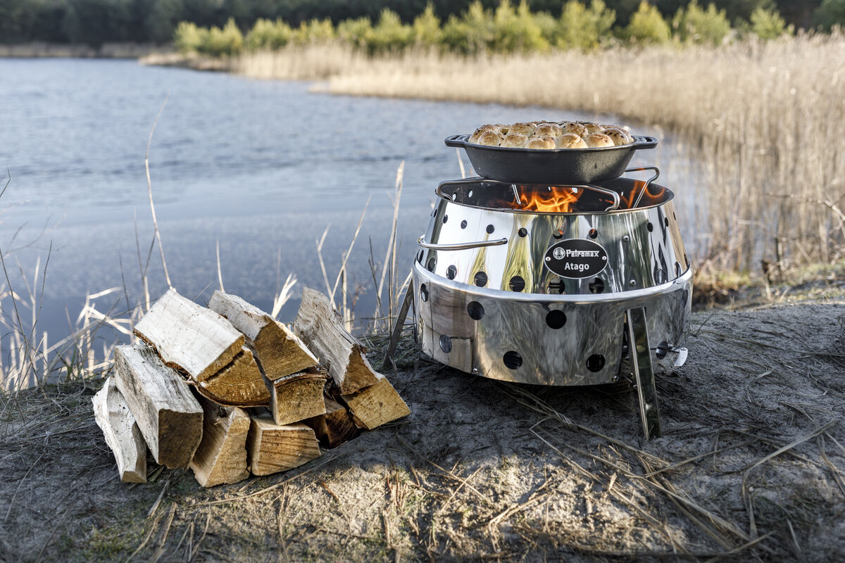 Ein runder, silberner Grill »Atago« auf einer natürlichen Fläche vor einem See. Der Grill brennt und eine Pfanne ist darüber platziert. Daneben liegt ein Stapel Holzscheite. Im Hintergrund sind Bäume und Schilf zu sehen.