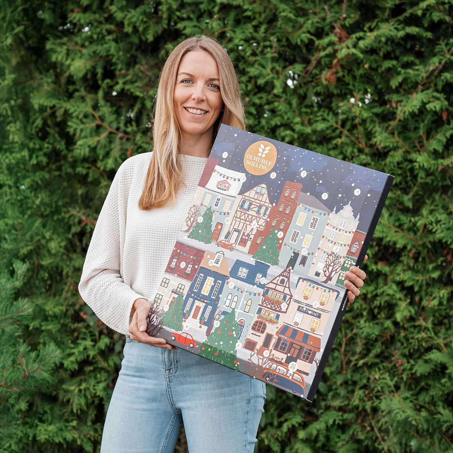 Eine Frau steht im Freien vor grünen Tannenbäumen und hält einen großen Adventskalender mit einer illustrierten Winterstadt in den Händen. Der Kalender zeigt bunte Häuser, verschneite Dächer und einen nachtblauen Himmel mit fallenden Schneeflocken. Das Motiv strahlt festliche und winterliche Stimmung aus.