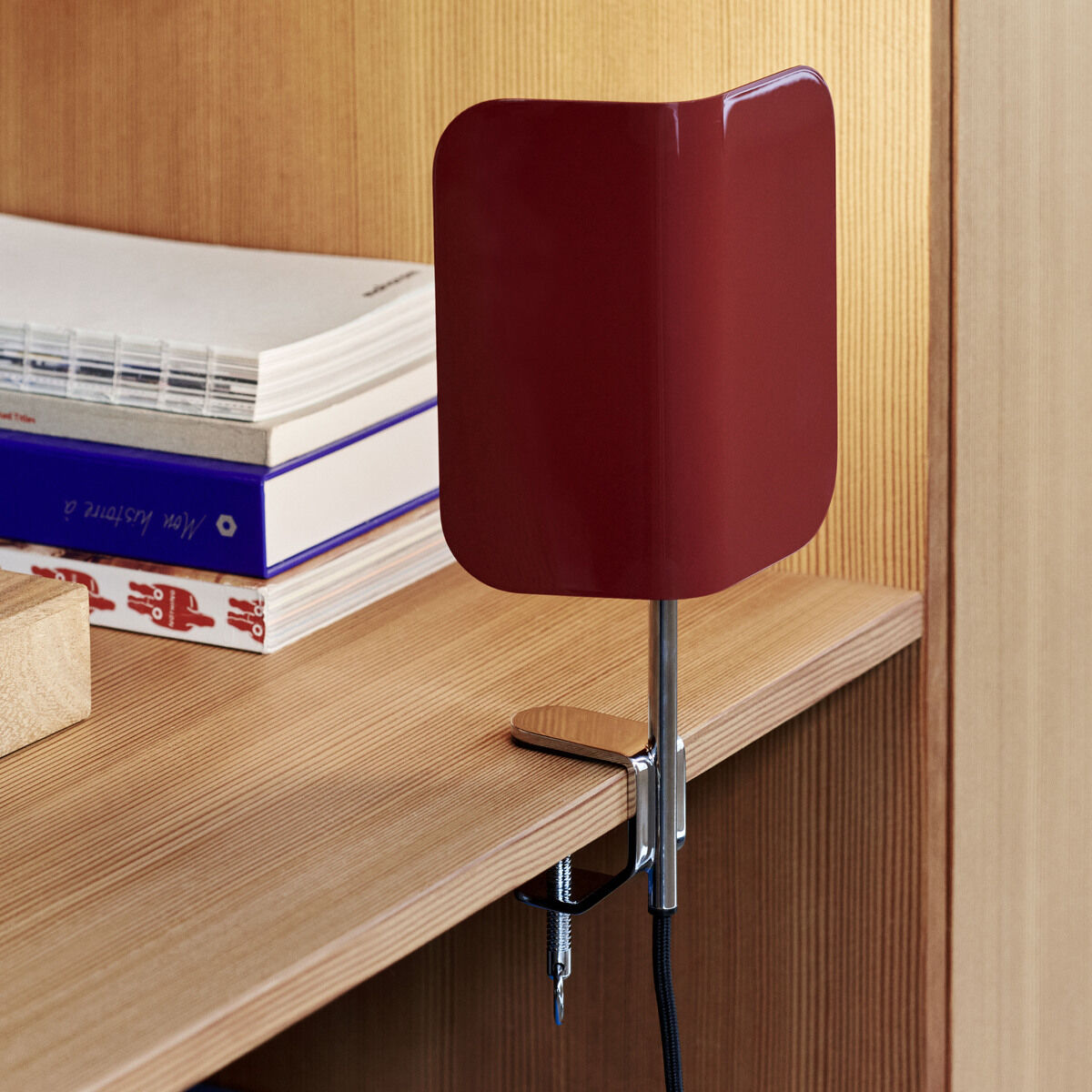 Rote Klemmlampe »Clip« von Apex an einem hölzernen Schreibtisch befestigt, mit zwei Schrauben und einem Kabel, umgeben von Büchern und anderen Gegenständen. Die Lampe hat ein minimalistisches, modernes Design mit einem silbernen Arm.