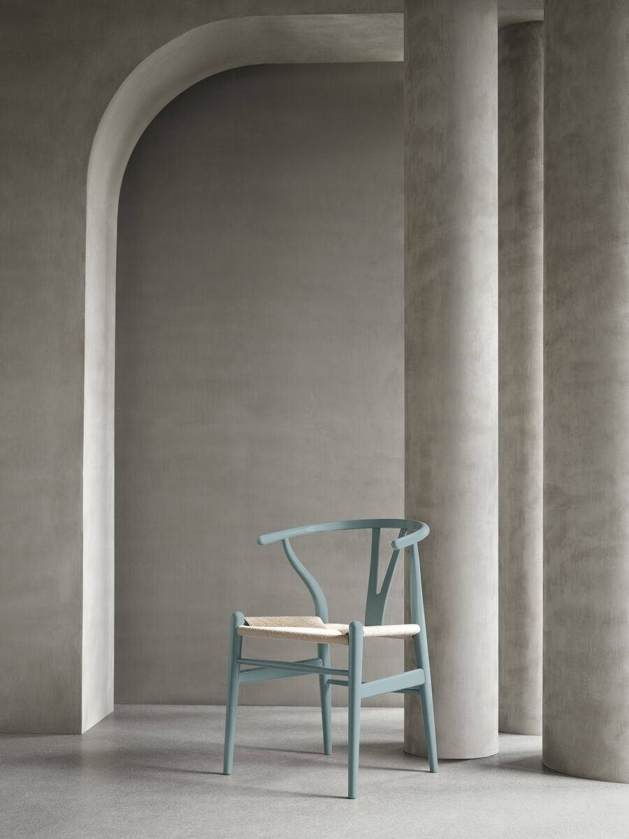 Ein moderner Stuhl, Modell Stuhl CH24 Soft von Carl Hansen, steht vor einer eleganten grauen Wand mit Bogendurchgänge. Der Stuhl hat eine blaue Lackierung mit einer naturfarbenen Sitzfläche. Minimalistisches skandinavisches Design.