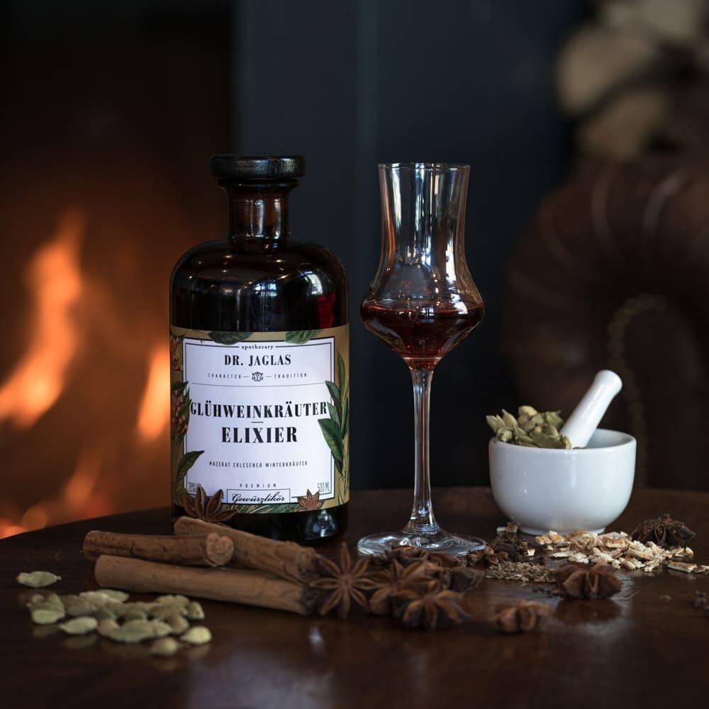Eine Flasche »Glühweinkräuter-Elixier« (Gewürzlikör) von Dr. Jaglas steht auf einem Holztisch. Daneben befindet sich ein elegant geformtes Likörglas, gefüllt mit dem Elixier. Verschiedene Gewürze wie Zimtstangen, Sternanis und Kardamom sind auf dem Tisch verstreut. Im Hintergrund ist ein unscharfes, warm erleuchtetes Ambiente zu sehen.
