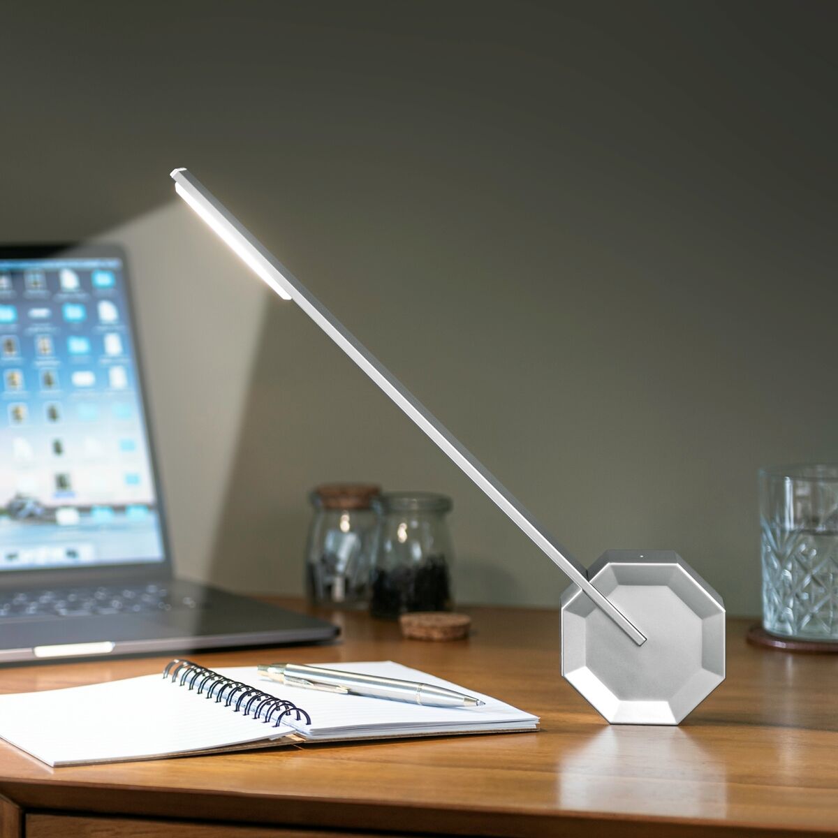 Octagon Tischlampe »One« auf einem Holztisch neben einem Laptop, Notizbuch und Stiften. Die Lampe hat ein modernes, minimalistisches Design mit einem schlanken, silbernen Rahmen und leuchtet hell in einem ansonsten gedämpften Raum.