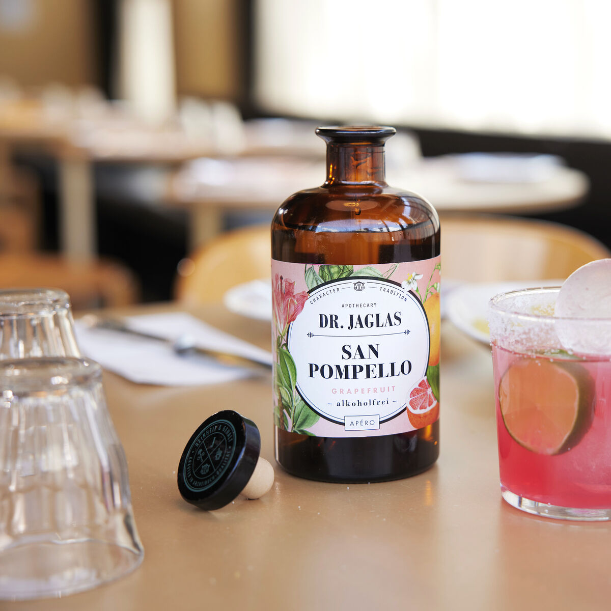 Flasche des alkoholfreien Aperitifs »San Pomello« von Dr. Jaglas neben einem Glas mit rosa Getränk und Limettenscheibe auf einem Tisch in einem Restaurantumfeld.