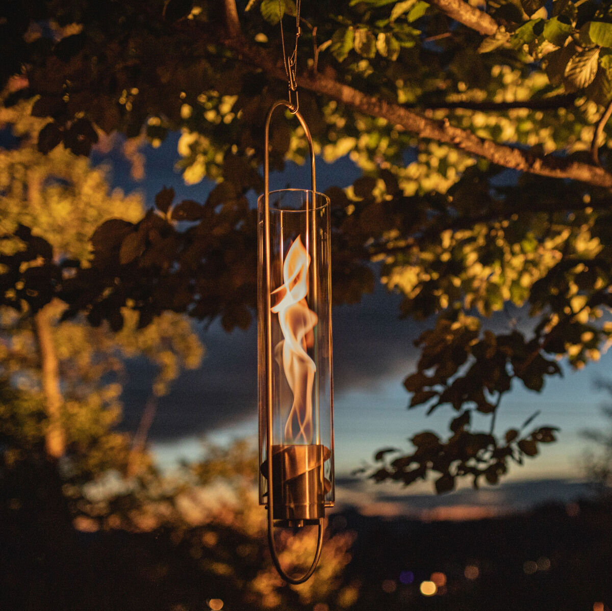 Ein SPIN Hängesystem hängt an einem Baum im Freien mit einem goldenen Feuerwirbel innen. Die Beleuchtung schafft eine gemütliche Atmosphäre in einem dunklen Garten mit wenig Licht, umgeben von grünen Blättern.
