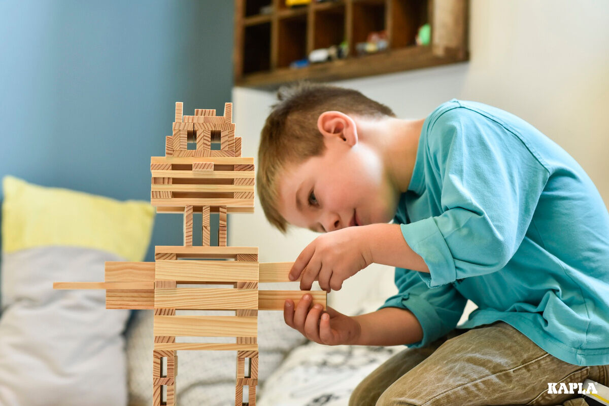 Ein Junge trägt ein blaues Oberteil und spielt im Kinderzimmer mit Holzbausteinen, indem er eine strukturierte Figur zusammensetzt. Im Hintergrund sind helle Kissen und weitere Spielzeuge zu sehen.