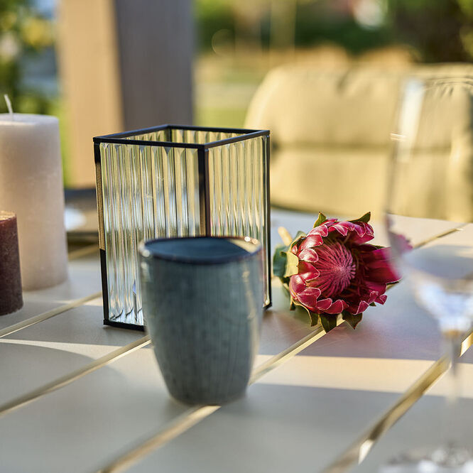 Ein gedeckter Tisch mit eleganter Dekoration, darunter eine Vase mit roter Blume, zwei Weinflaschen, Teelichtkerzen und Gläser auf einem modernen, beigen Holztisch in einem Außengartenbereich.