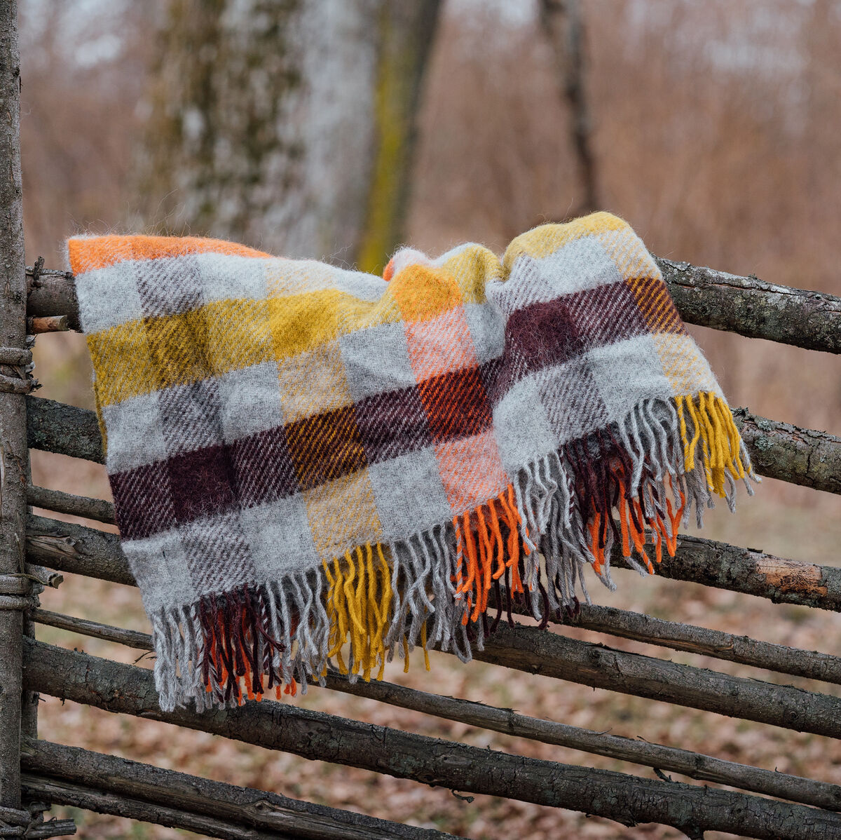 Eine handgewebte Decke im Karomuster, genannt Gotland Multi, liegt über einem rustikalen Holzzaun. Die Decke zeigt Herbstfarben wie Orange, Gelb, Rot, Braun, Grau und Weiß und hat Fransen an den Enden.