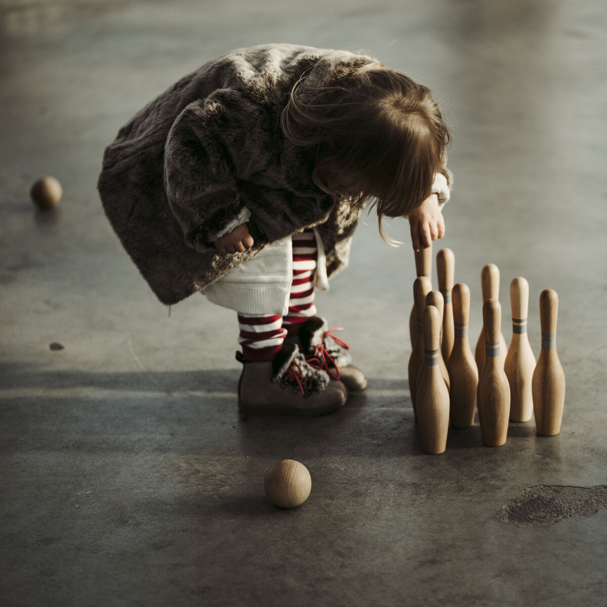 Ein Kind in einem Pelzmantel beugt sich über mehrere aufgestellte Holzkegel und eine Holzkugel auf einem Boden aus dunklem Beton, um den Kegelspiel zu spielen.