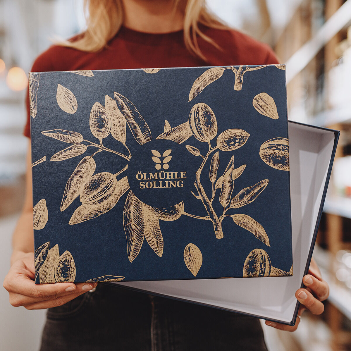 Eine Person hält eine geöffnete, exklusiv gestaltete ZEIT Präsentbox Jubiläumsessige von Ölmühle Solling. Die Box zeigt ein botanisches Muster mit gold-beigen Illustrationen auf dunkelblauem Grund sowie das Firmenlogo in der Mitte. Die Verpackung wirkt hochwertig und edel.