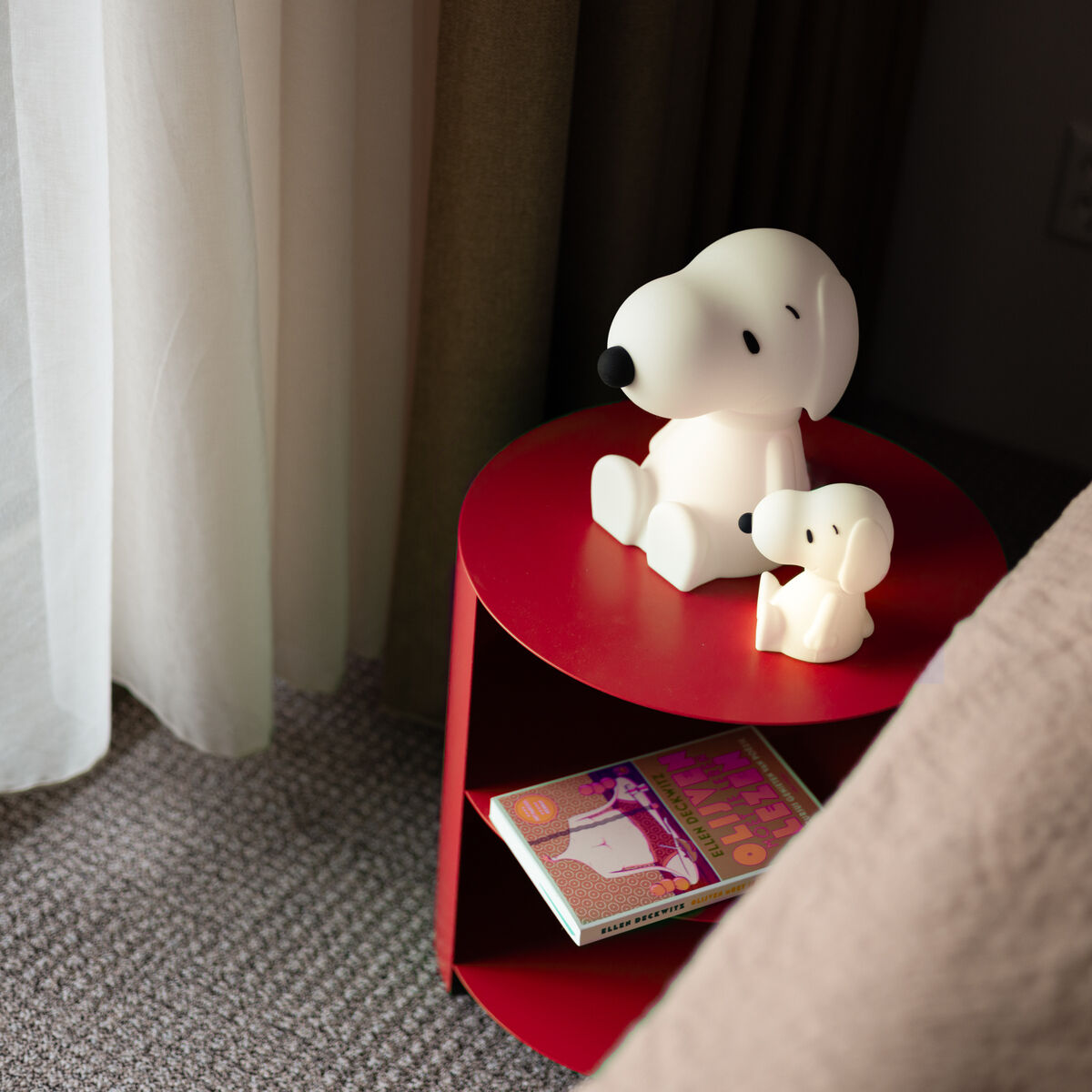 Auf einem roten Nachttisch stehen zwei Snoopy-Nachtlichter in verschiedenen Größen neben einem Bett. Unter dem Tisch liegt ein bebildertes Kinderbuch. Durch das Fenster fällt Tageslicht ins Zimmer.