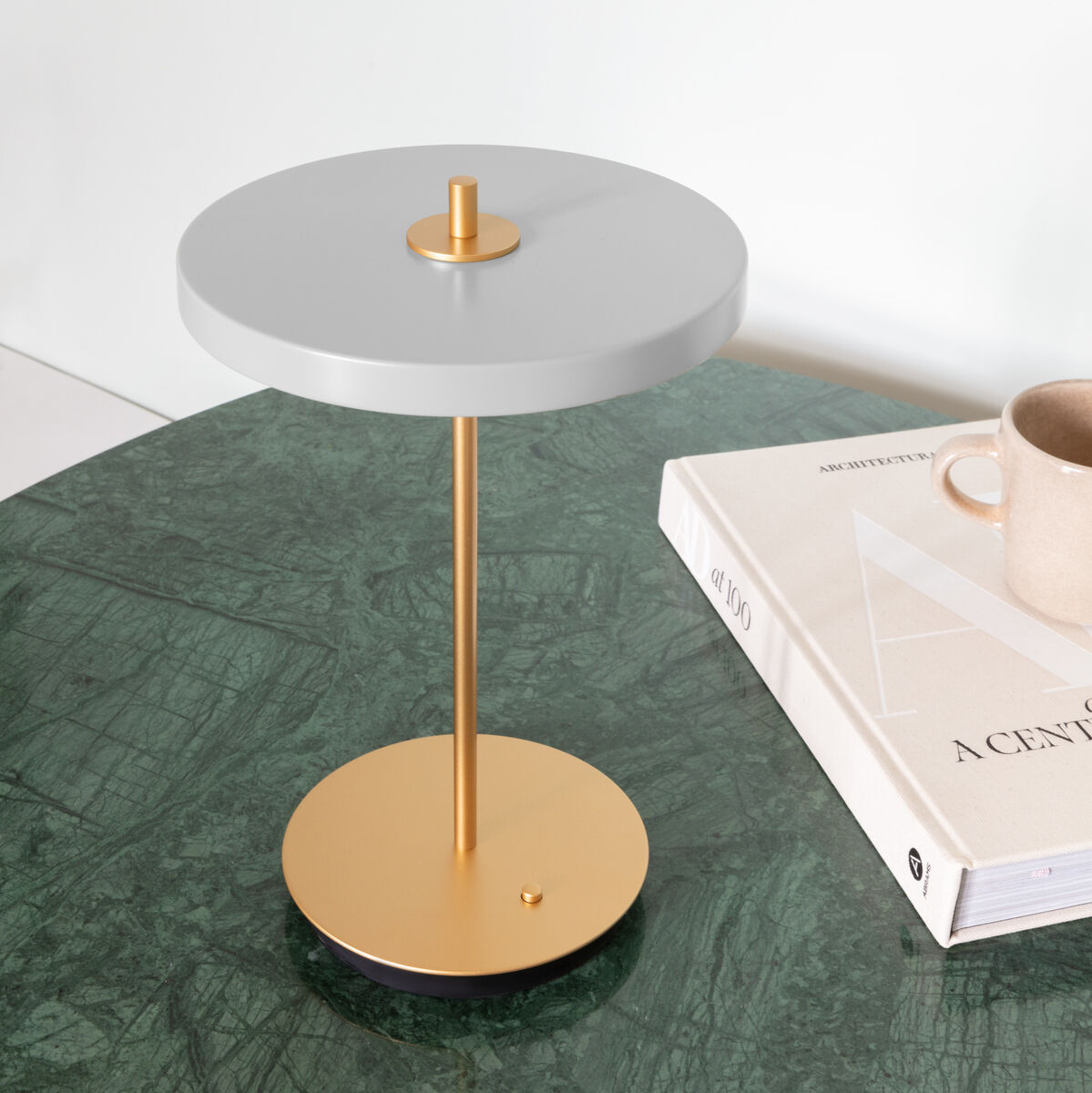 Elegante, moderne Tischlampe »Asteria Move II« mit goldener Basis und grauem Lampenschirm. Die Lampe steht auf einem grünen Tisch neben einem Buch und einer beige Tasse.