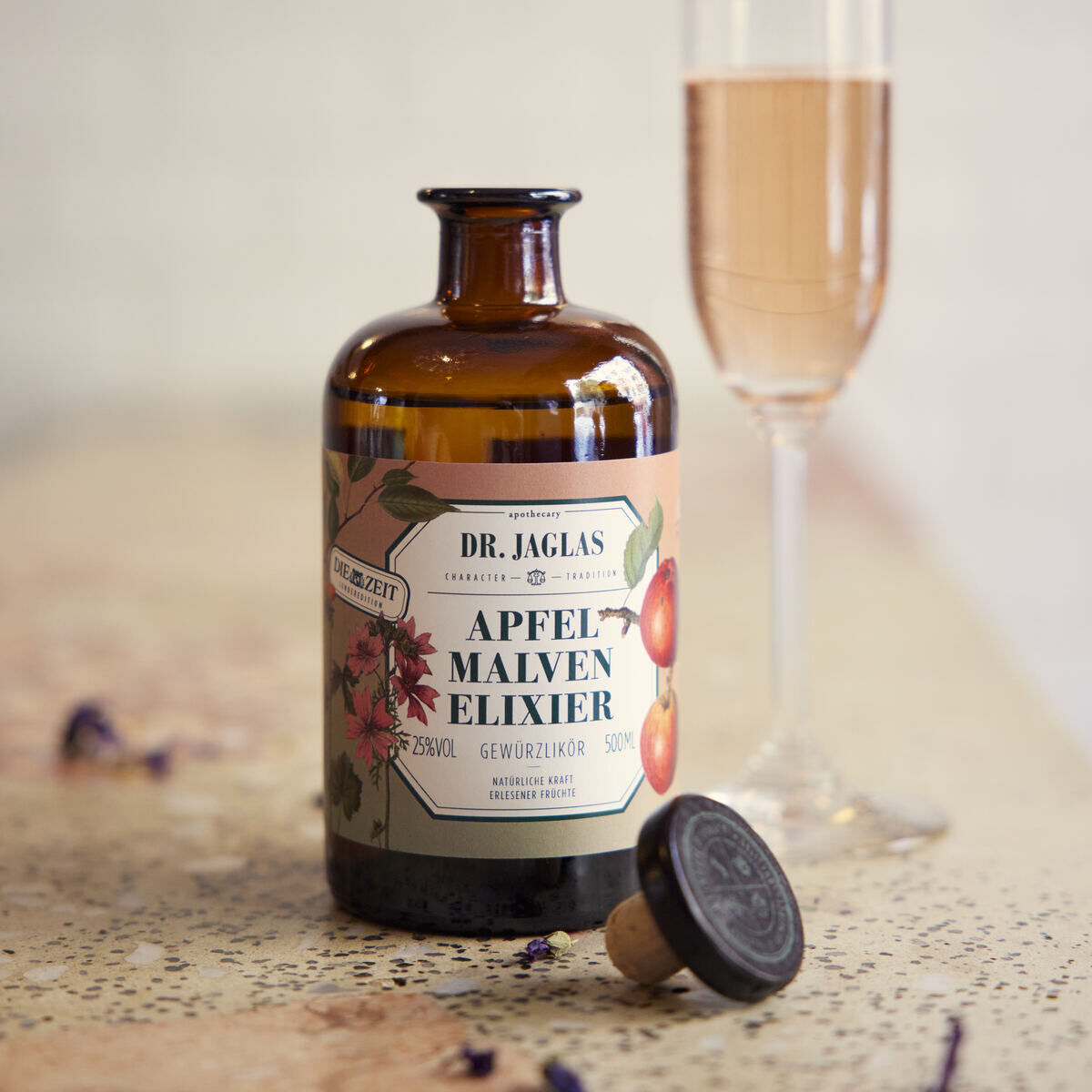 Eine Flasche Dr. Jaglas Apfel Malven-Elixier Dessertlikör steht neben einem halbgefüllten Sektglas auf einem mit Blumenblüten dekorierten Tisch. Der Korken der Flasche liegt neben der Flasche.