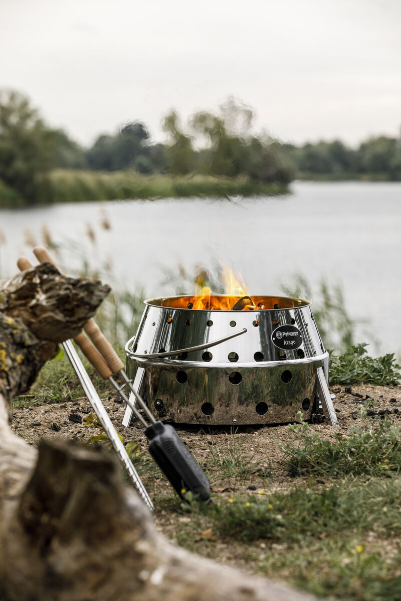 Grill »Atago« aus Edelstahl im Einsatz, aufgestellt am Ufer eines Sees mit brennendem Feuer, umgeben von Natur und Campingzubehör.