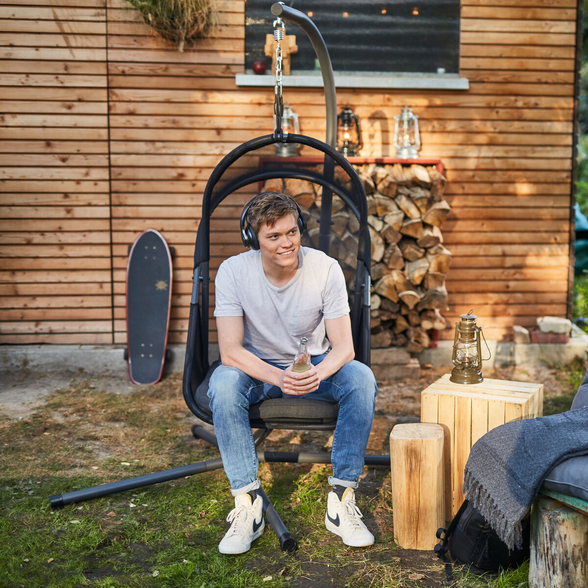 Ein Mann sitzt in einem schwarzen Hängesessel »Yobaya« im Garten neben einem kleinen Tisch und einer gepolsterten Bank. Im Hintergrund ist eine Holzwand sowie einige dekorative Elemente wie Laternen und gestapeltes Holz zu sehen. Der Fokus liegt auf einer entspannten Atmosphäre im Freien.