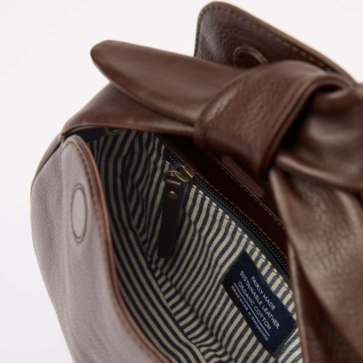 Naheaufnahme einer braunen Ledertasche mit Schleifendetail am Verschluss. Das geöffnete Innenfach zeigt ein gestreiftes Stofffutter in creme und dunkelblau sowie ein eingenähtes Markenetikett und einen Reißverschluss. Die Tasche wirkt elegant und modern und eignet sich für den täglichen Gebrauch.