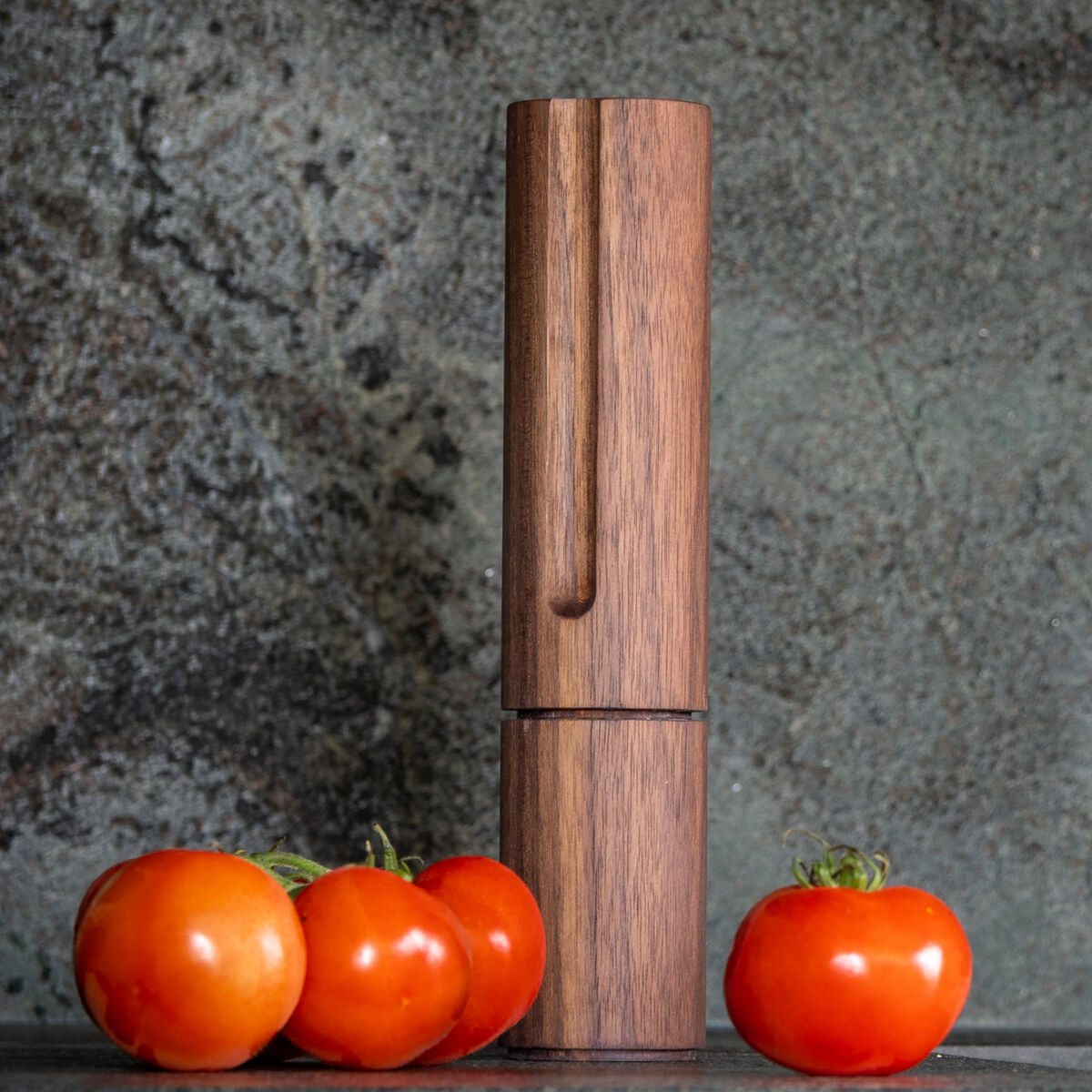 Auf einer modernen, grauen Küchenarbeitsfläche stehen die Gewürzmühle »TURN« aus Walnussholz von finbeaux und vier rote Tomaten. Rechts befindet sich ein moderner schwarzer Wasserhahn. Natürliche Materialien und minimalistisches Design prägen das Bild.