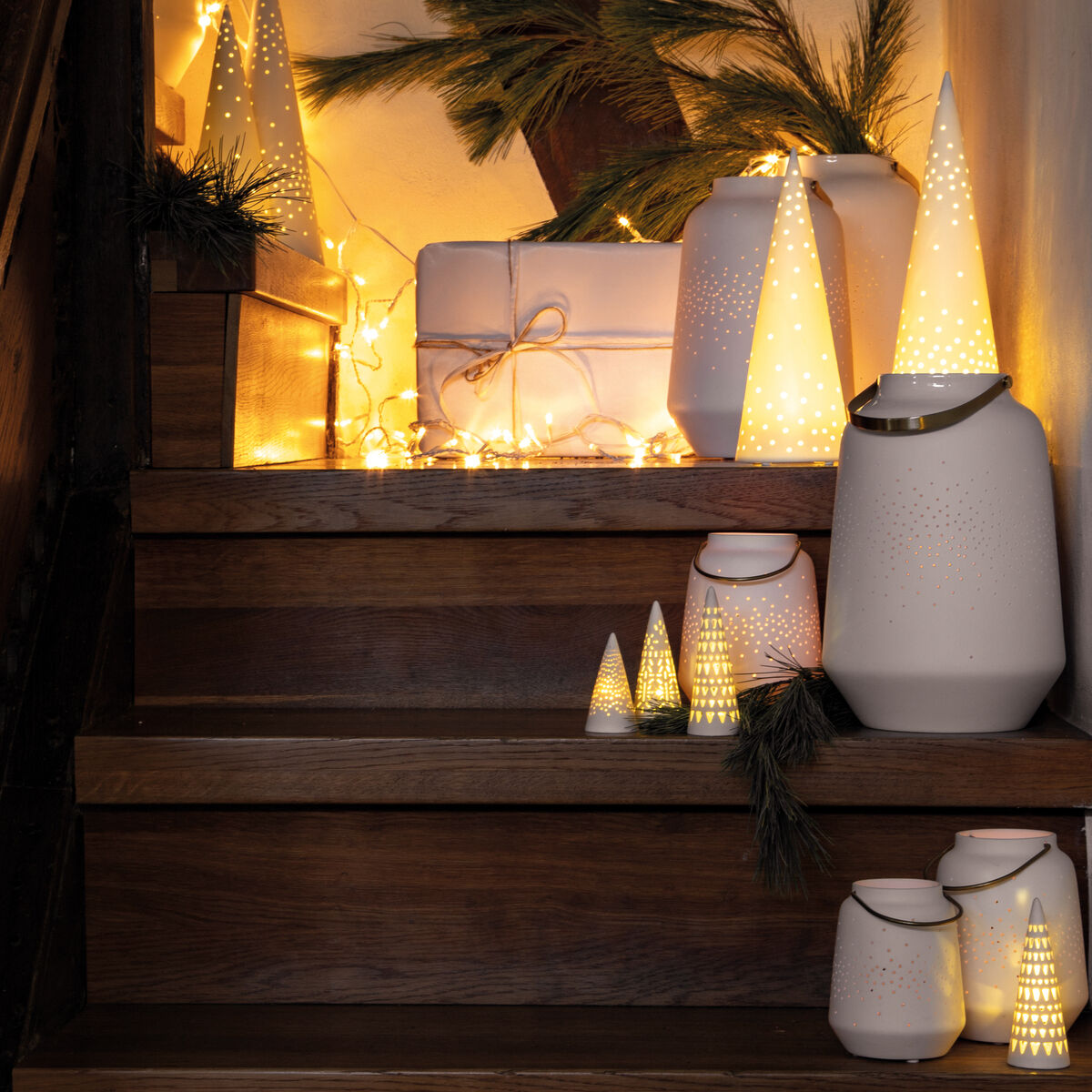 Eine stimmungsvolle Weihnachtsdekoration auf einer Holztreppe mit Porzellanlaternen, Lichterketten und Geschenken. Die Beleuchtung ist warm und gemütlich, akzentuiert durch Tannenzweige.