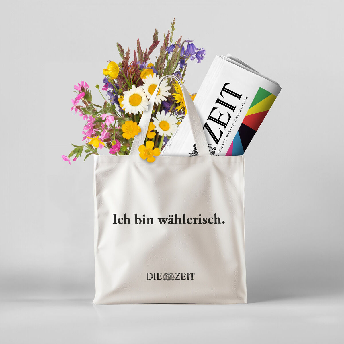 Eine weiße Stofftasche mit der Aufschrift 'Ich bin wählerisch' und einem Blumenstrauß sowie einer Zeitung. Der Hintergrund ist grau und schlicht.