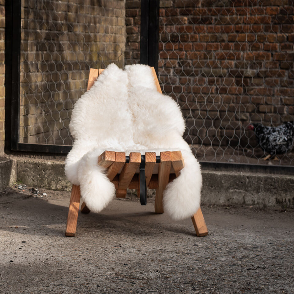 Ein flauschiges weißes Schafsfell mit Lederschlaufe liegt dekorativ auf einem modernen Holzstuhl im Außenbereich; im Hintergrund eine Ziegelmauer und ein Huhn.