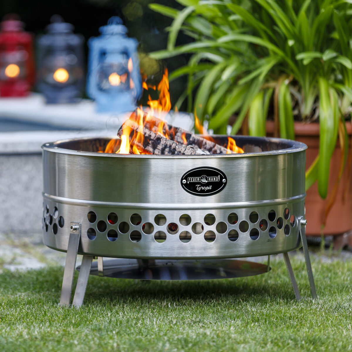 Ein Bild zeigt eine »Tyropit« Feuerschale im Garten. Die Schale ist aus Metall mit Lüftungslöchern und steht auf einem Rasen. Brennholz liegt daneben. Im Hintergrund leuchten bunte Laternen, und eine Pflanze im Topf ist sichtbar.
