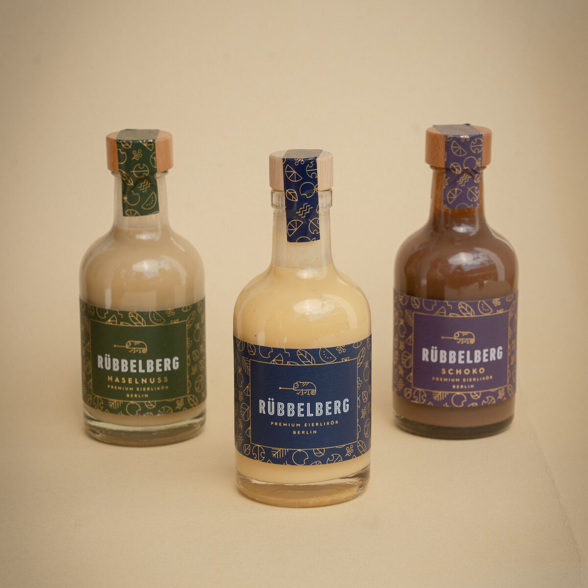 Das Bild zeigt drei 200ml-Flaschen Premium Eierlikör von Rübbelberg in verschiedenen Sorten. Jede Flasche hat ein individuelles farbiges Etikett und einen Naturholzkorken. Die Flaschen stehen nebeneinander vor einem hellen Hintergrund. Geeignet als edles Geschenk oder für besondere Genussmomente.