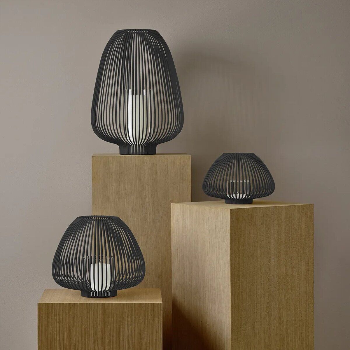 Drei moderne Laternen aus dunklem Metall in unterschiedlichen Größen stehen auf hellen Holzsäulen vor einer beigen Wand. Jede Laterne enthält ein weißes Windlicht und schafft eine stilvolle, minimalistische Wohnatmosphäre.