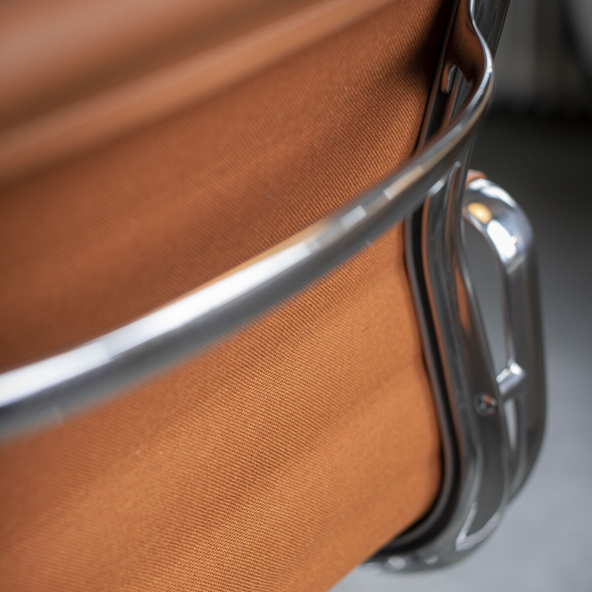Nahaufnahme eines Vitra Alu Chair EA 118 in einer polierten ZEIT-Sonderedition. Der Stuhl hat einen braunen Stoffbezug und metallische Elemente aus poliertem Aluminium, die einen glänzenden und stilvollen Look erzeugen. Der Boden ist dunkelgrau.