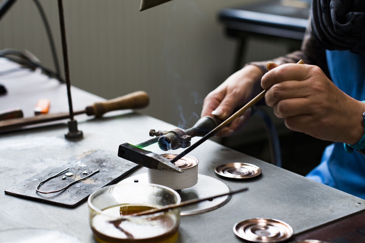 Eine Person arbeitet an einem Metallschmiedetisch und hantiert mit Werkzeugen. Auf dem Tisch befinden sich verschiedene metallische Gegenstände und ein Glas mit einer Flüssigkeit.