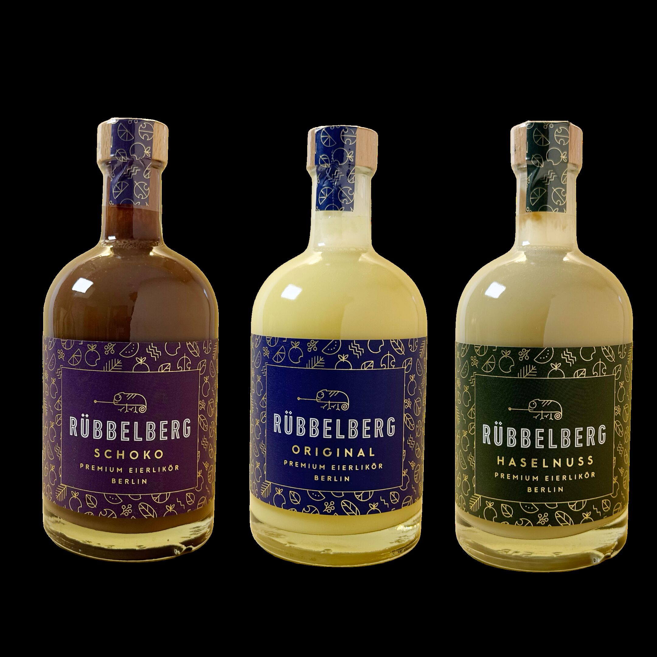 Drei 500ml-Flaschen Premium Eierlikör der Marke Rübbellberg stehen nebeneinander. Die Varianten sind Schoko, Original und Haselnuss, jeweils mit einem individuell gestalteten Etikett in Dunkelviolett, Creme und Dunkelgrün. Die Flaschen sind transparent und zeigen unterschiedlich gefärbte Liköre. Das Produkt wirkt hochwertig und ist als Geschenkset geeignet.