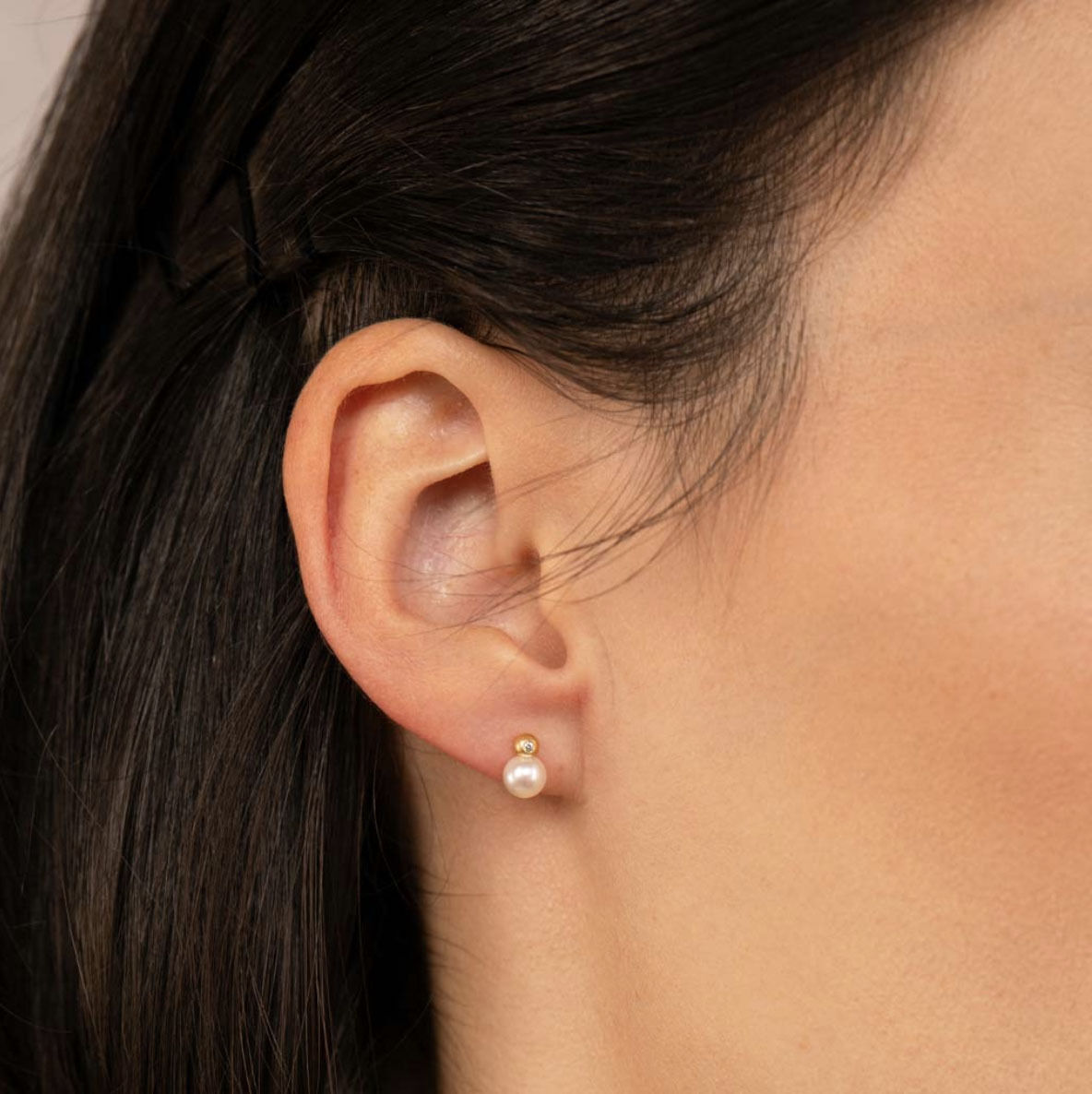 Detailaufnahme eines weiblichen Ohrs mit dem Ohrstecker Star von Eva Strepp Schmuckdesign. Der Stecker besteht aus vergoldetem Silber mit einer runden weißen Süßwasserzuchtperle. Dezentes, elegantes Design, Hintergrund und Haare in dunklen, neutralen Tönen.