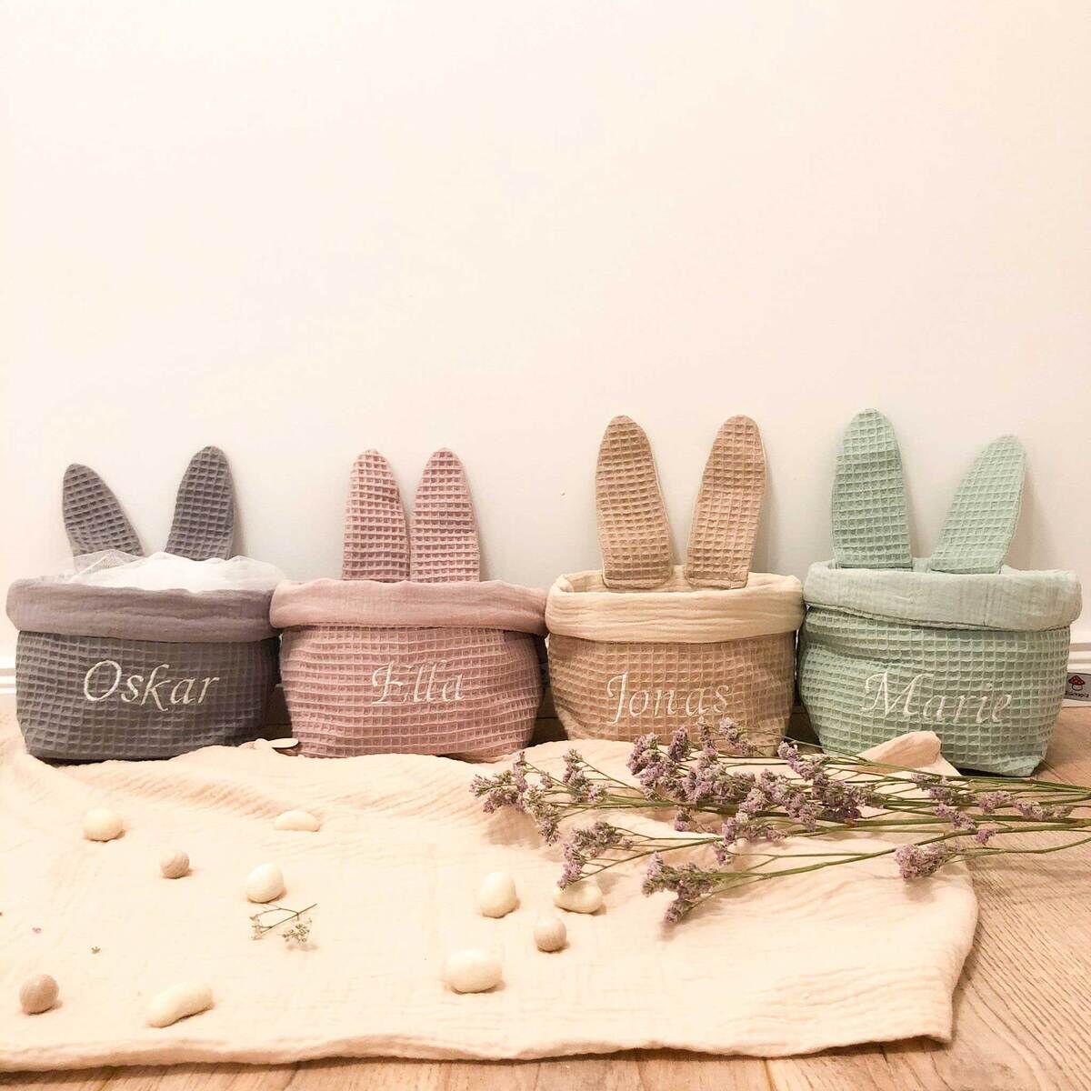 Vier Osterkörbchen in Hasenform in den Farben Grau, Rosa, Beige und Mintgrün, die jeweils mit Namen bestickt sind, stehen auf einem Tuch mit blassen Blüten und kleinen Steinen.