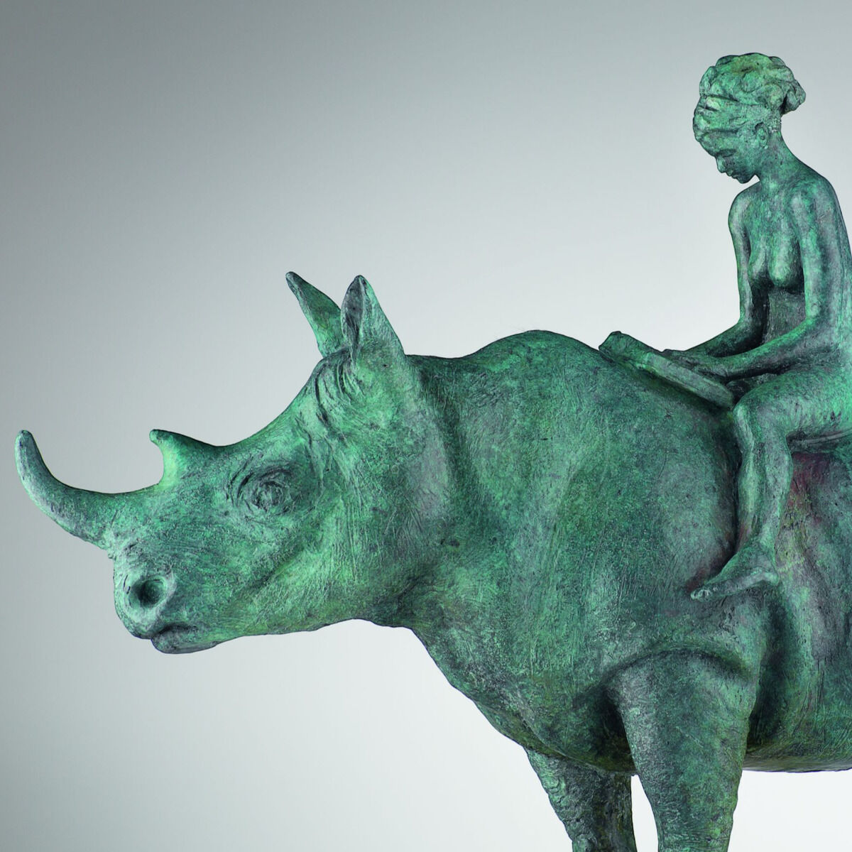 Seitliche Produktaufnahme der Bronzeskulptur „The Maid and the Unicorn“ von Alfredo Llorens García: Ein kräftiges Nashorn mit langem Vorderhorn trägt eine kleine, aufrecht sitzende lesende Frauenfigur auf dem Rücken. Die rau strukturierte, grün-türkis patinierte Bronze steht auf einem schmalen weißen Sockel vor hellgrauem Hintergrund.