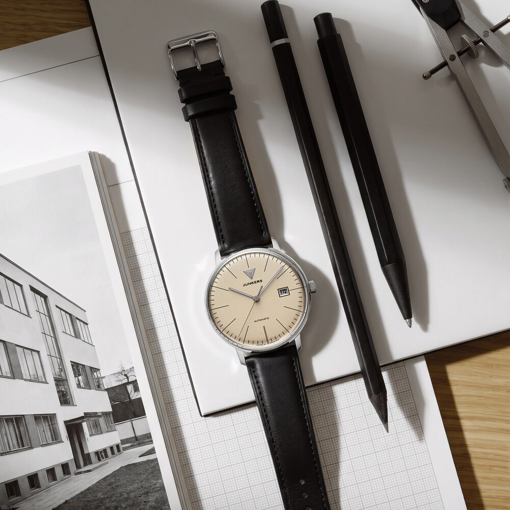 Die Junkers ZEIT-Sonderedition »Bauhaus Automatik« liegt mit schwarzem Lederarmband und beigem Zifferblatt auf kariertem Papier. Daneben liegen schwarze Stifte und ein Zirkel, im Hintergrund ein Bauhaus-Foto. Die Szenerie vermittelt schlichte, funktionale Eleganz.