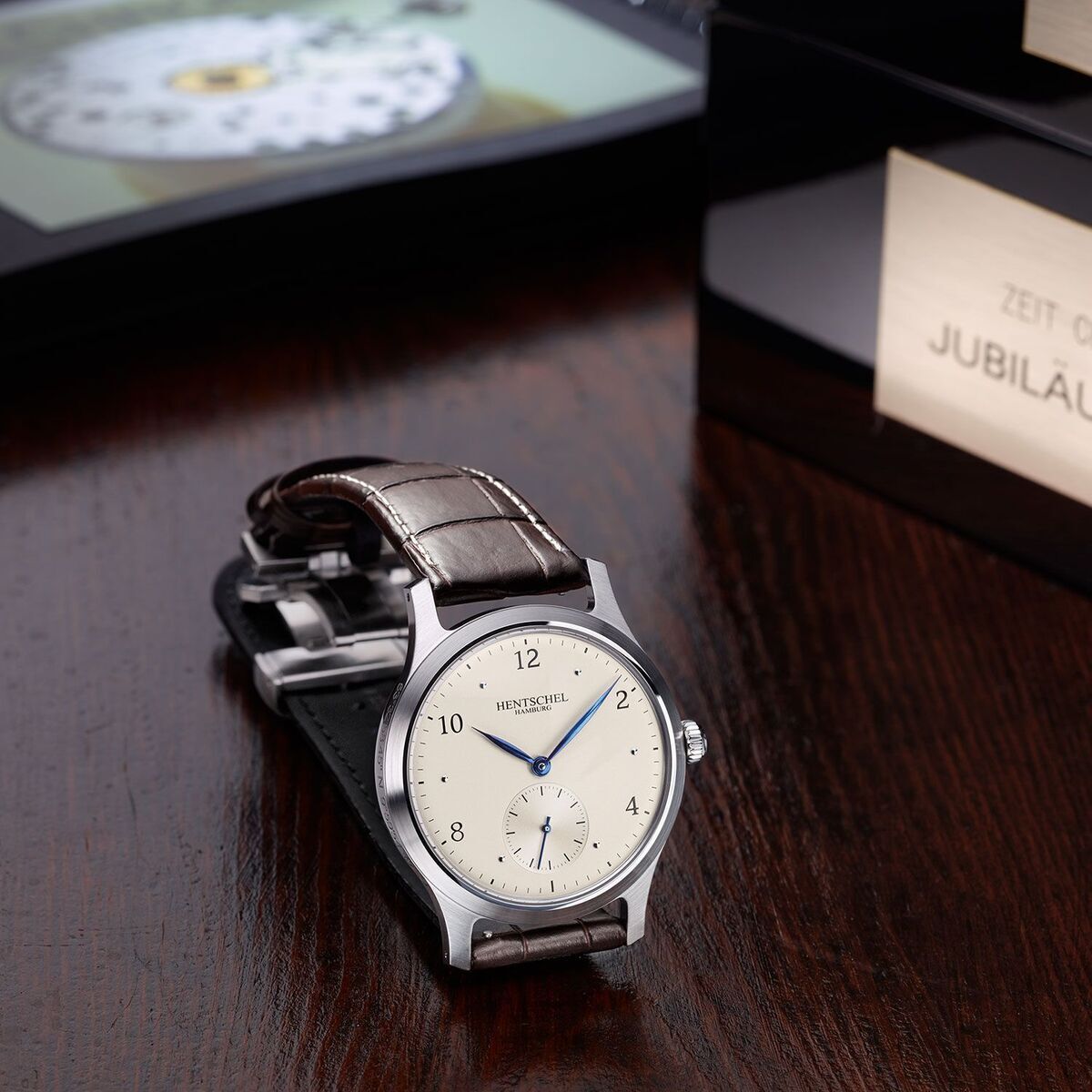 Eine elegante Herrenuhr der Marke ZEIT-Chronometer, Modell »H2 Hamburg«, mit einem klassischen Design. Sie verfügt über ein silbernes Gehäuse, ein weißes Zifferblatt und ein braunes Lederarmband. Die Uhr liegt auf einer dunklen Holzoberfläche neben einer Geschenkschachtel mit der Aufschrift 'Jubiläumsedition'.