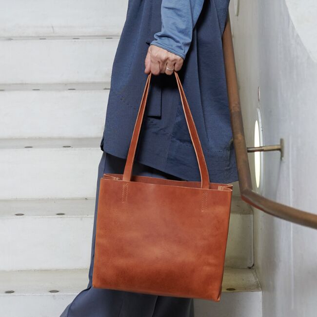 Eine moderne Paperbag-Tasche aus cognacfarbenem Leder wird von einer Person an der Seite getragen. Die Tasche präsentiert sich in schlichtem, minimalistischem Design mit zwei Tragegriffen und passt ideal zu einem eleganten oder lässigen Outfit. Das Foto ist auf einer weißen Treppe mit rundem Fenster im Hintergrund aufgenommen und zeigt nur einen Ausschnitt der Person mit Fokus auf das Accessoire.