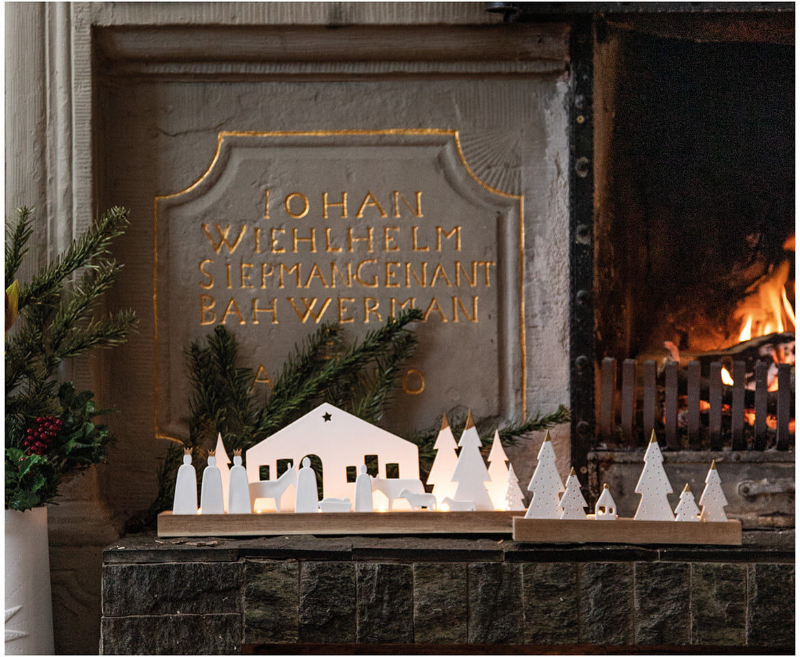 Große Holzkrippe auf einem Kaminsims, umgeben von Tannenzweigen, vor einem dekorativen Hintergrund mit Inschrift und neben einem brennenden Kamin.