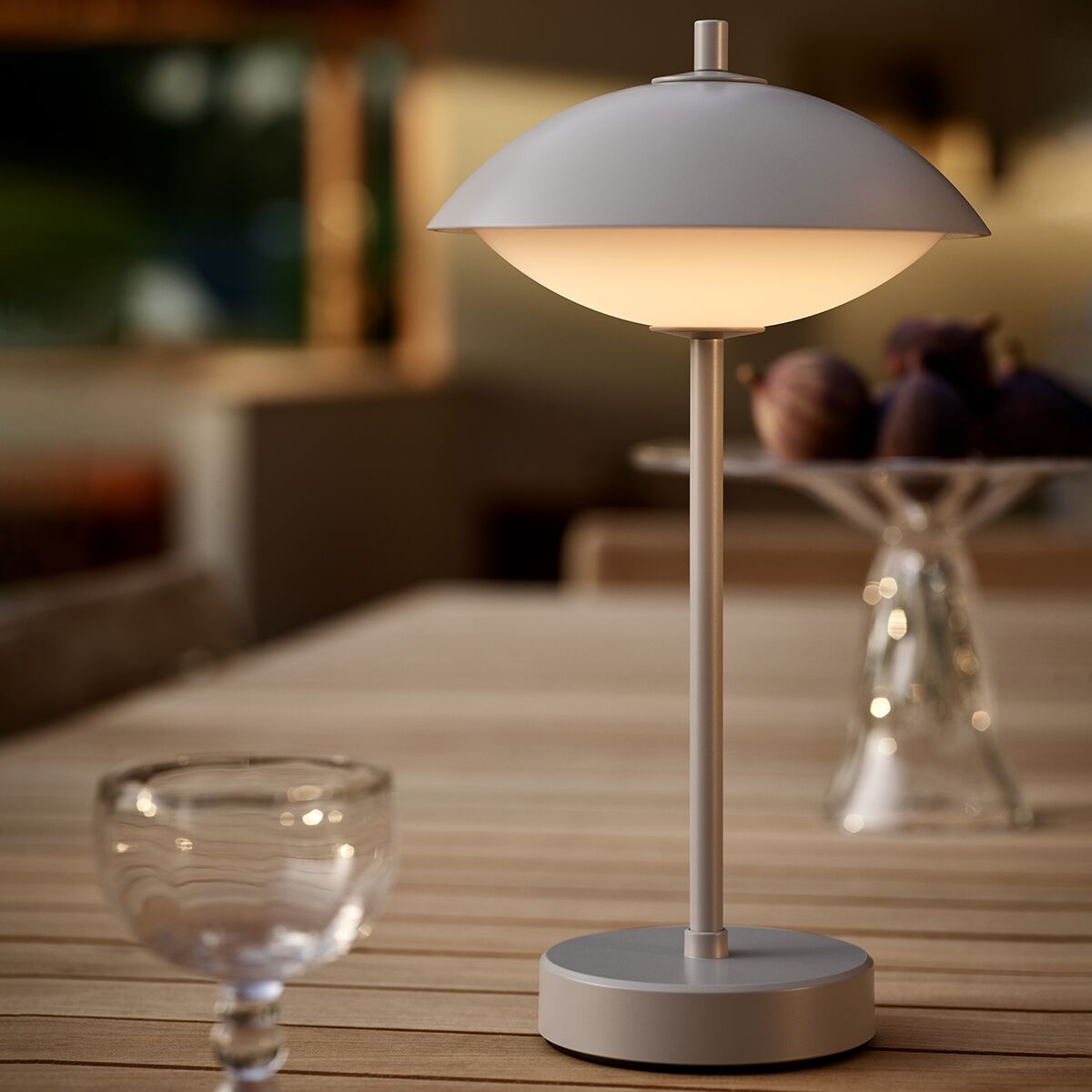 Die tragbare Lampe »Clam™« von Fritz Hansen steht eingeschaltet auf einem hellen Holztisch in einem modern eingerichteten Wohnraum. Die Lampe hat einen pilzförmigen, weißen Schirm auf einem schlanken, grauen Standfuß. Im Hintergrund unscharfe Deko und ein Glas im Vordergrund.