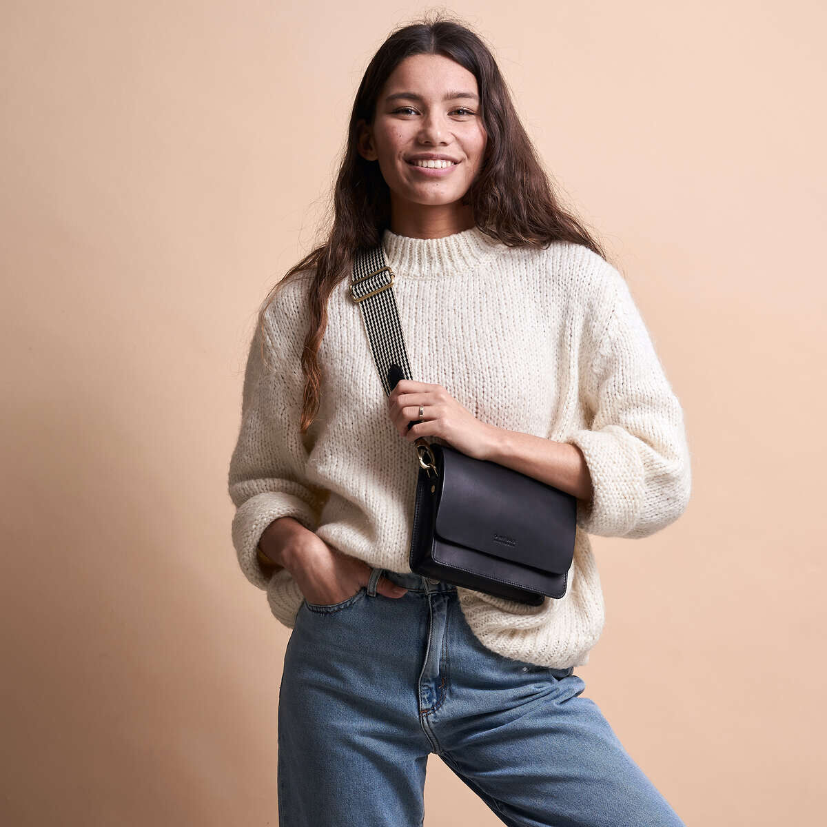 Junge Frau mit einer schwarzen Tasche »Audrey Mini« in einer lässigen Pose mit einem cremefarbenen Pullover und blauen Jeans vor einem beigefarbenen Hintergrund.
