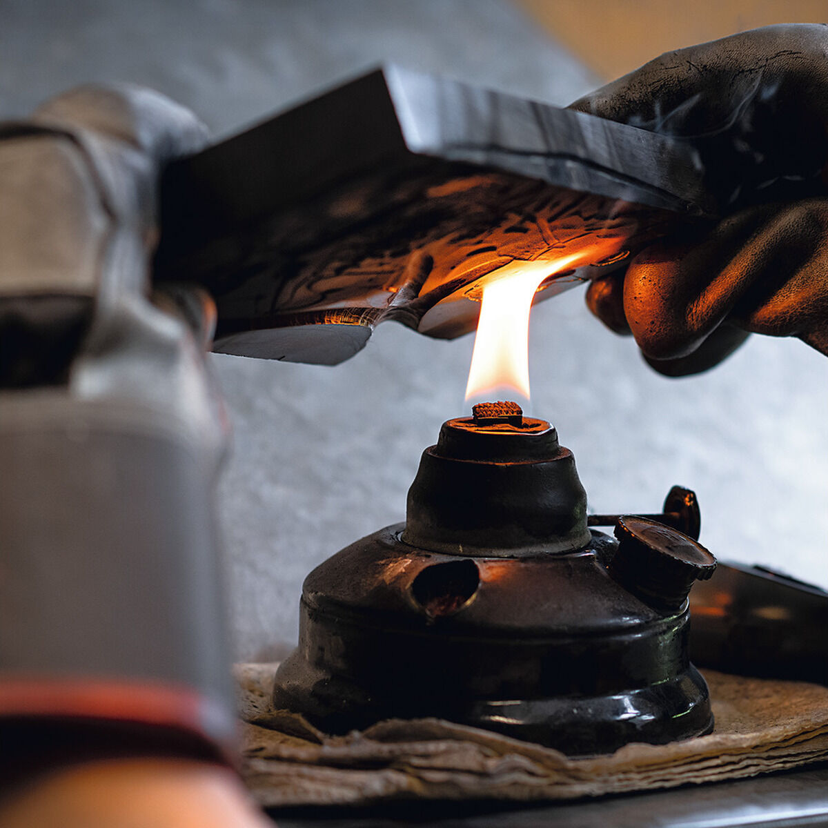 Detailaufnahme eines Handwerkprozesses in einer Werkstatt: Eine Person mit Schutzhandschuhen hält eine Metallform über eine offene Flamme. Materialien wie Zinn und Metall sind erkennbar, im Hintergrund ein grauer Tisch. Die Szene wirkt industriell und präzise.