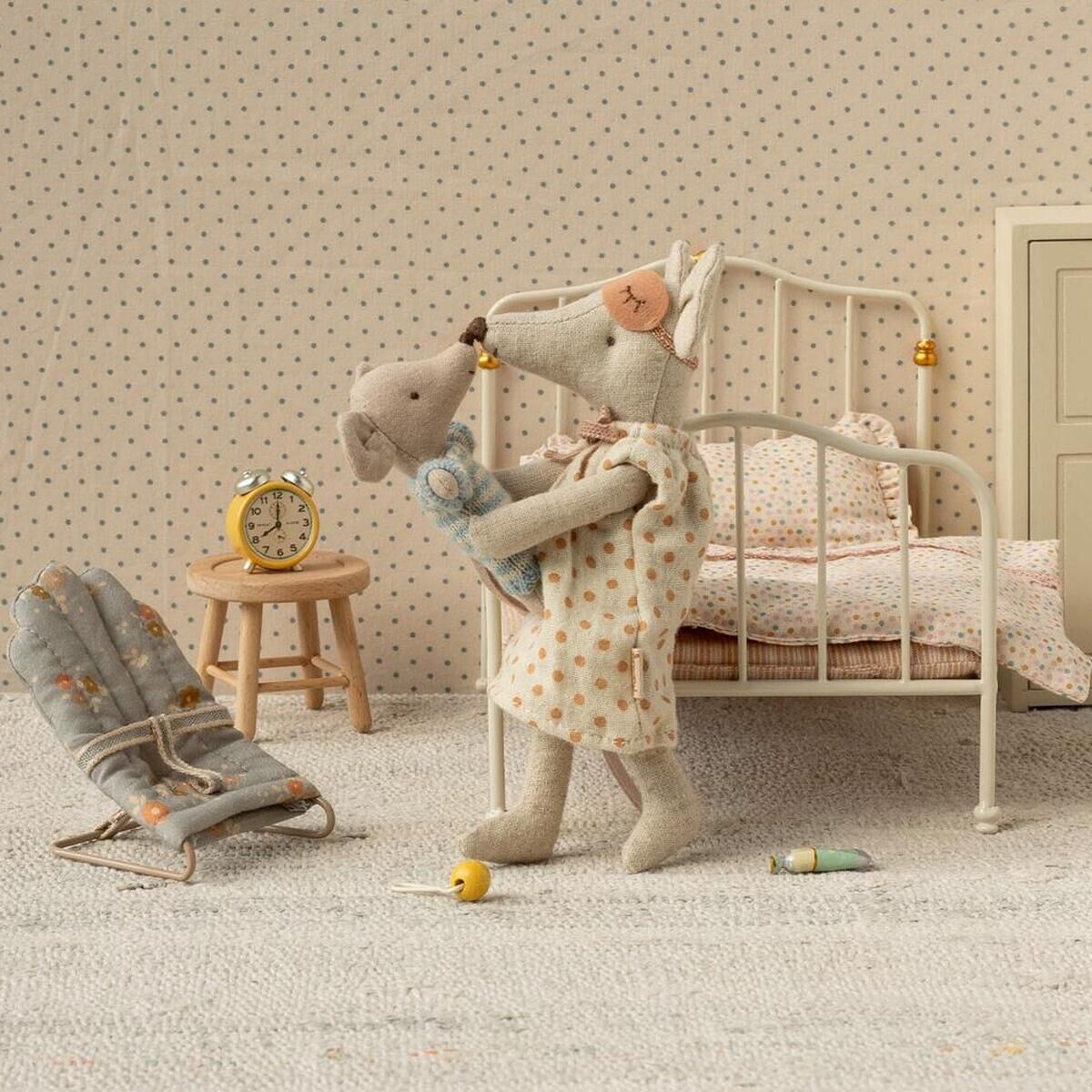 Zwei Stoffmäuse, dargestellt als Mama und Papa, stehen in einem detailreich eingerichteten Miniatur-Kinderzimmer. Die Szene zeigt ein Metallbett, einen kleinen Nachttisch mit Wecker, einen Babystuhl und abgestimmte, dezente Farben. Die Mäuse halten ein Stoffbaby im Arm und wirken liebevoll und fürsorglich.