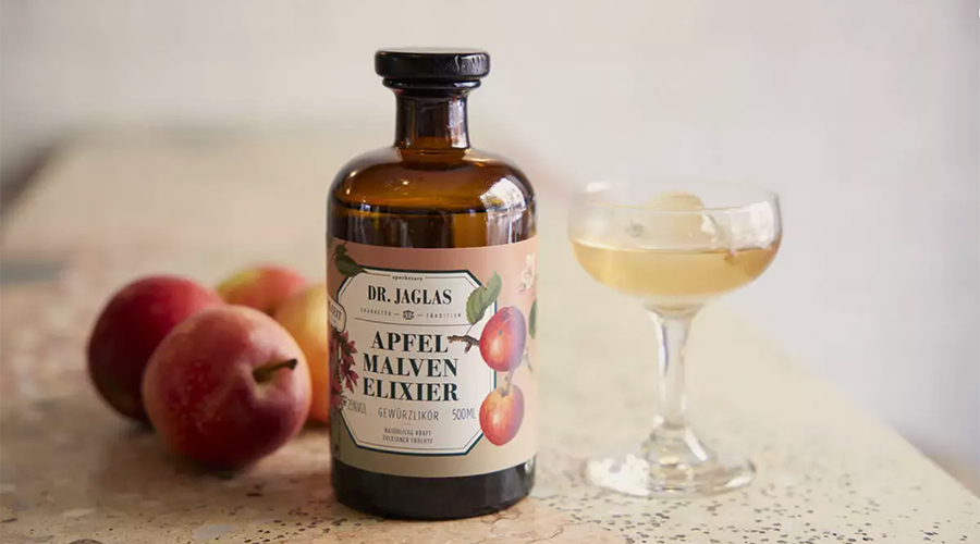 Braune Flasche Dr. Jaglas Apfel Malven Elixier Likör neben Glas mit Eiswürfeln und zwei roten Äpfeln auf Tisch